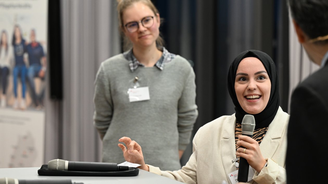 Eine junge Frau steht am Tisch und beantwortet die Frage eines Moderators, im Hintergrund eine andere junge Frau