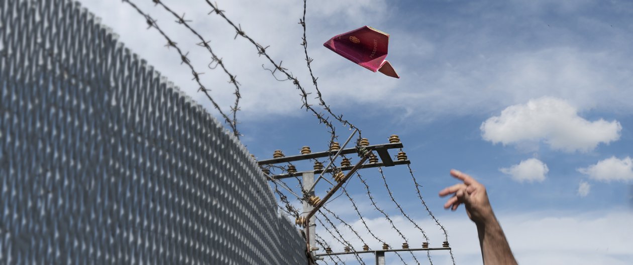A paper airplane flies over a border fence.
