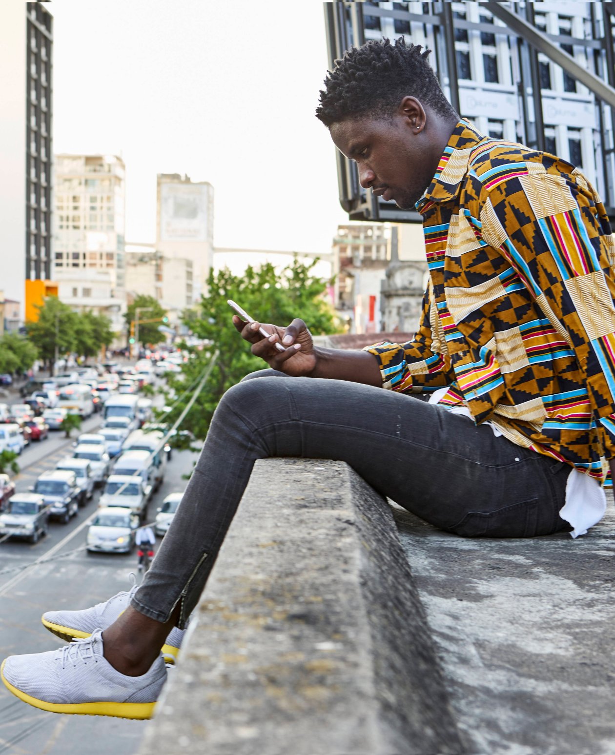 Young man sits on a wall in an African city and looks into his cell phone.