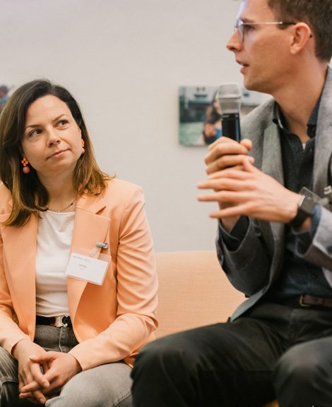 Two people are sitting at a table in front of a laptop, having a conversation.