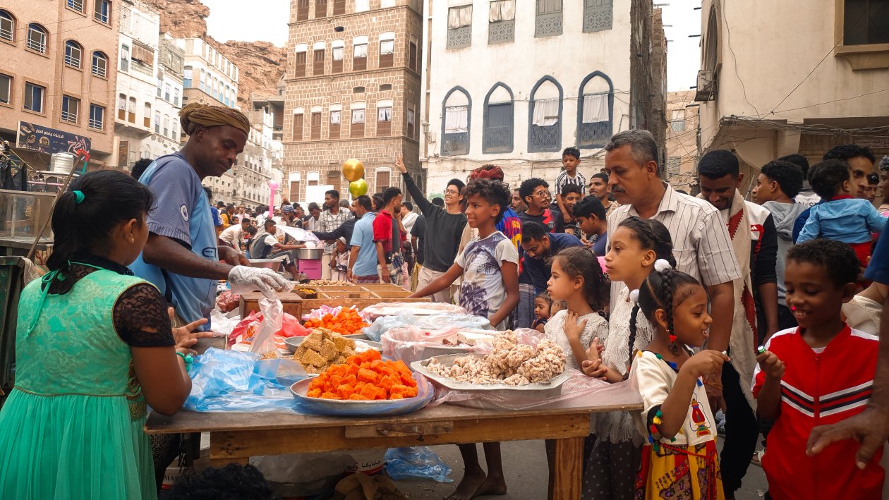 Ein Markstand inmitten von Menschen: ein Mann bietet einigen KIndern und einem Vater in Mukalla Essen an.