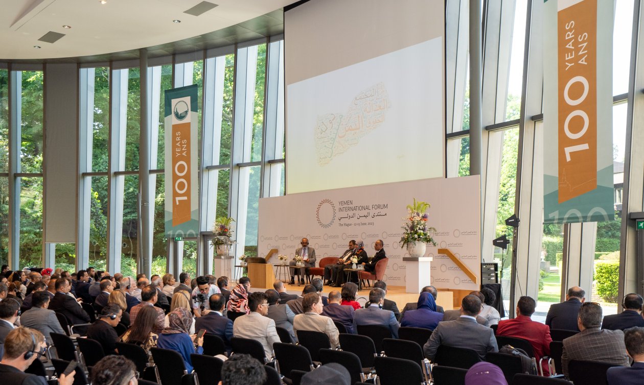 Crowd sitting in a huge forum room facing a glass front and a podium, whre four people are debating