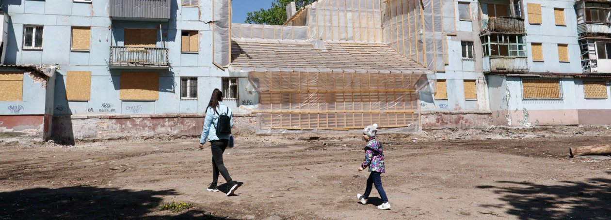 Mutter und Tochter laufen über einen Platz, im Hintergrund zerstörte Gebäude mit Baugerüsten