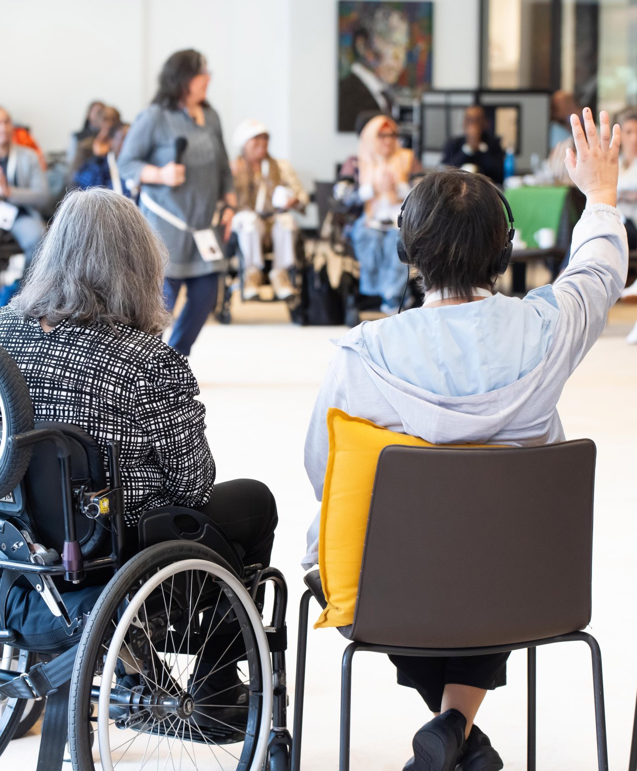 People in a discussion group. In the foreground, a person in a wheelchair; another person raises their hand.