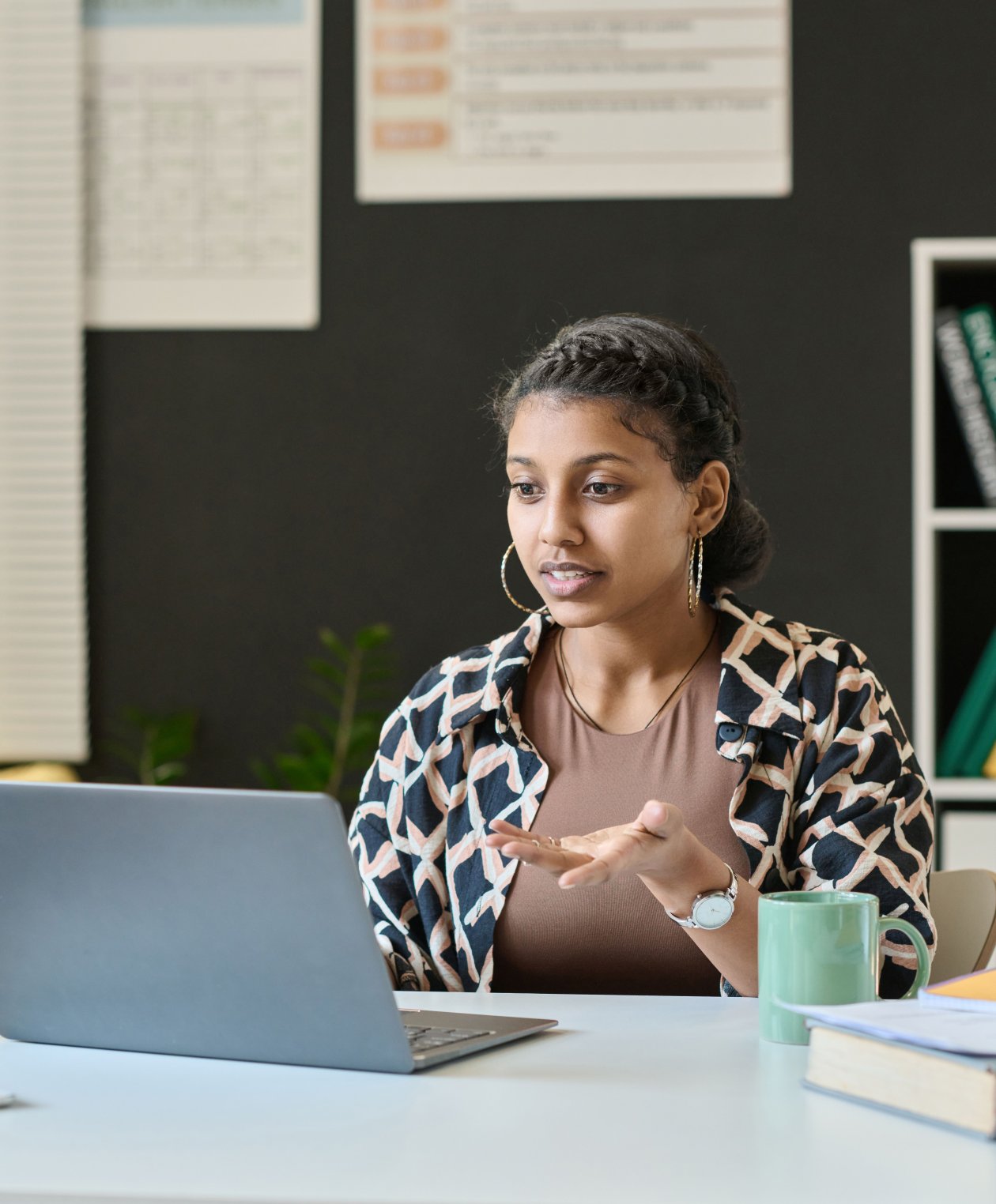 A student is sitting at desk in front of laptop and talking to teacher online on laptop