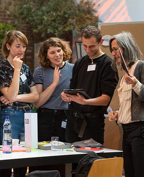 Four people are standing around a work table discussing something. In the background, there is a pinboard with notes on it.