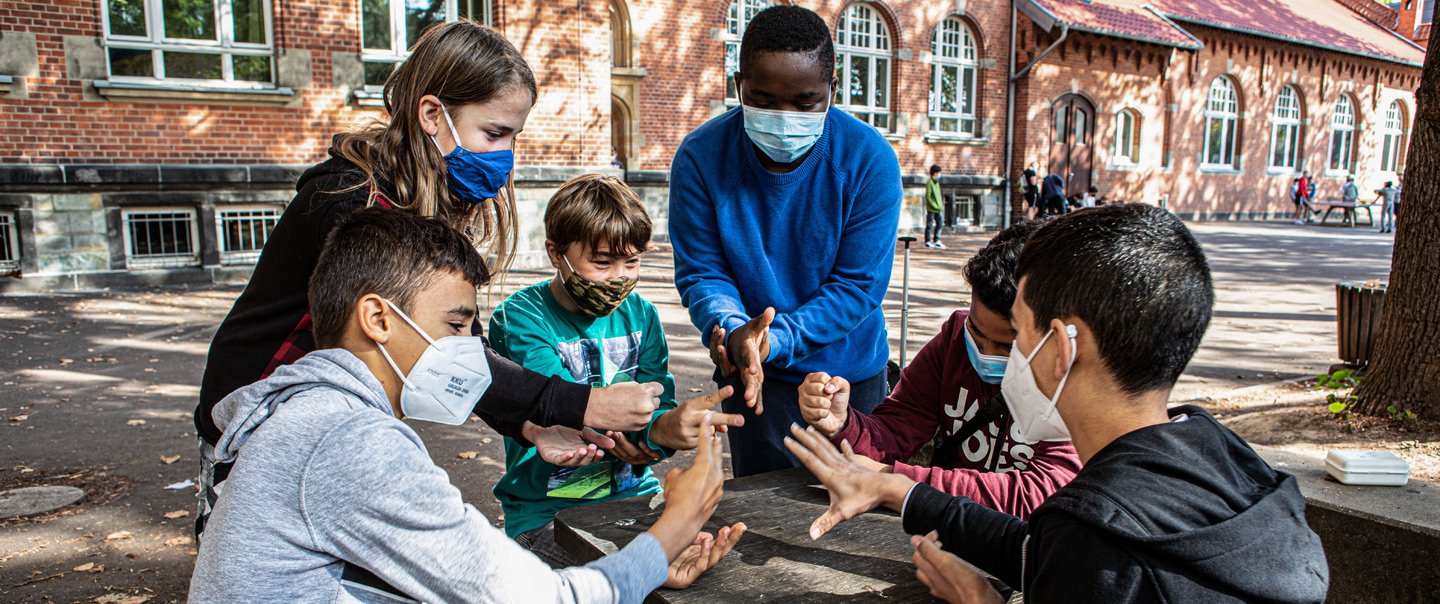 Sechs Schüler spielen auf dem Schulhof Schere, Stein, Papier. Sie tragen Mund-Nasen-Schutz.