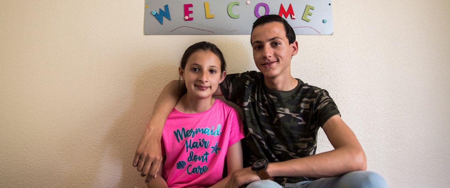 Two young people sit in front of a poster with the inscription &quot;Welcome&quot;.