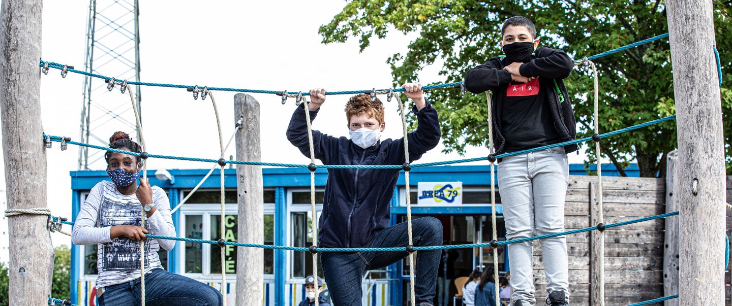 Ein Mädchen und zwei Jungen mit Masken stehen auf einem Klettergerüst.