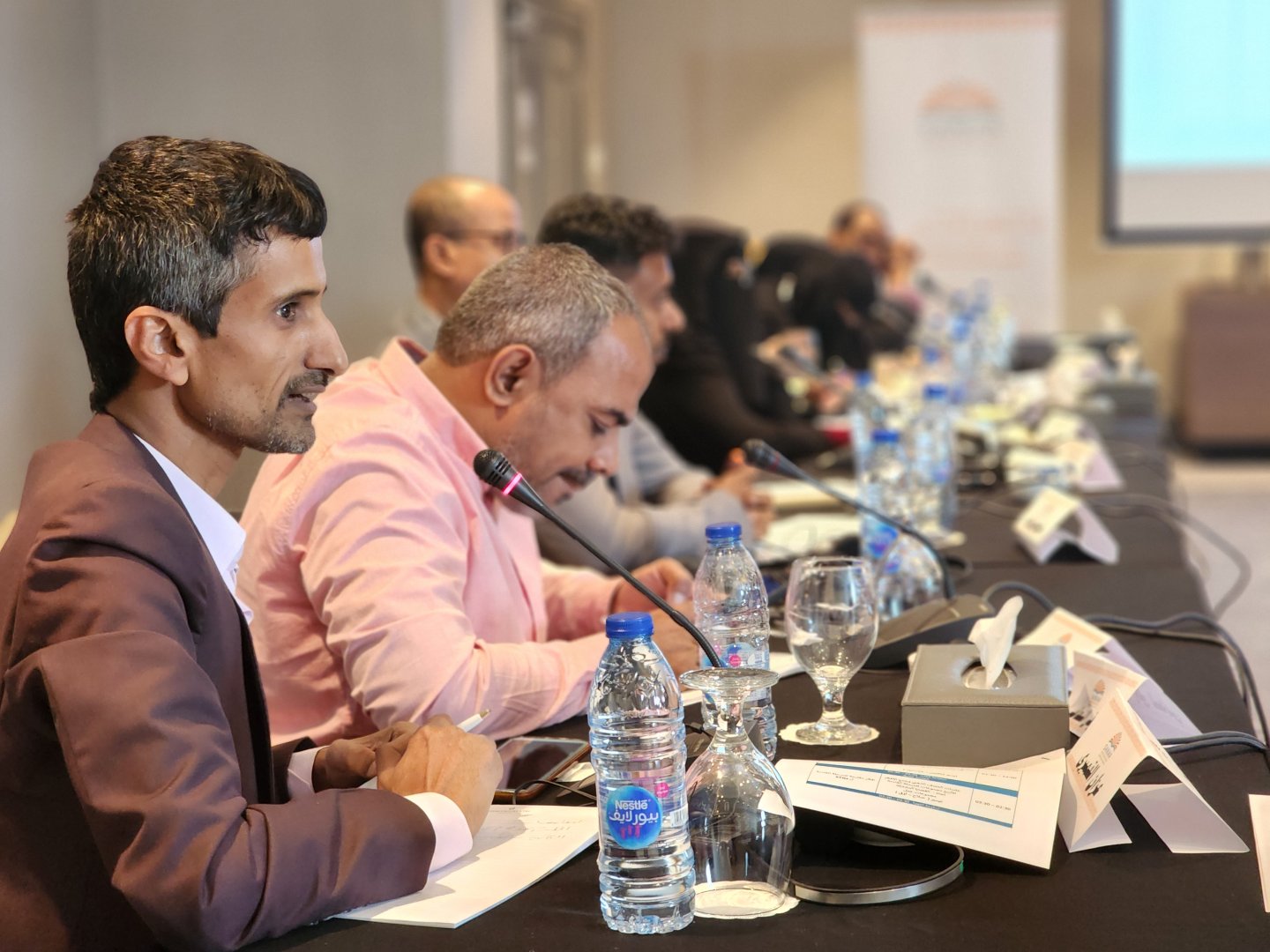 Sideview: Azzan Al-Qumairi, seated at a long table next to other persons lined up behind