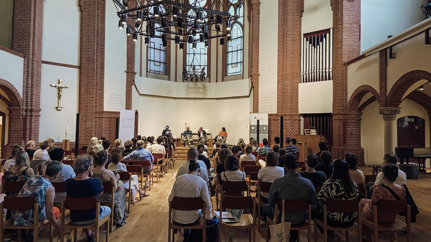 People in a church, sitting in rows and listening to a discussion that is seen in front