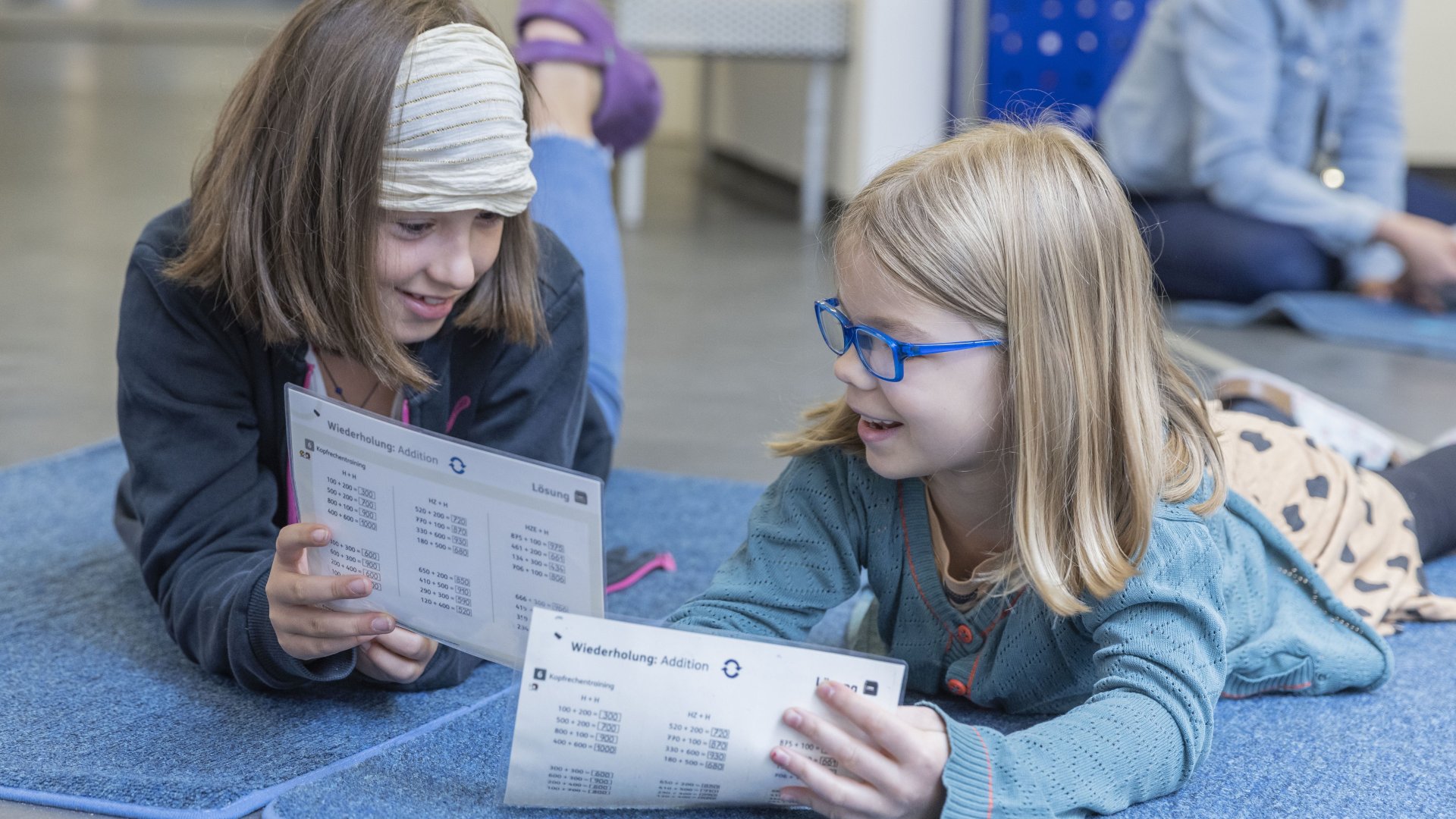 Two girls lie on the floor in a school, holding math exercises in their hands