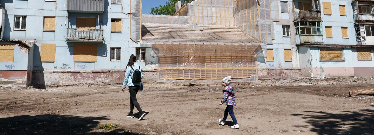 Mutter und Tochter laufen über einen Platz, im Hintergrund zerstörte Gebäude mit Baugerüsten