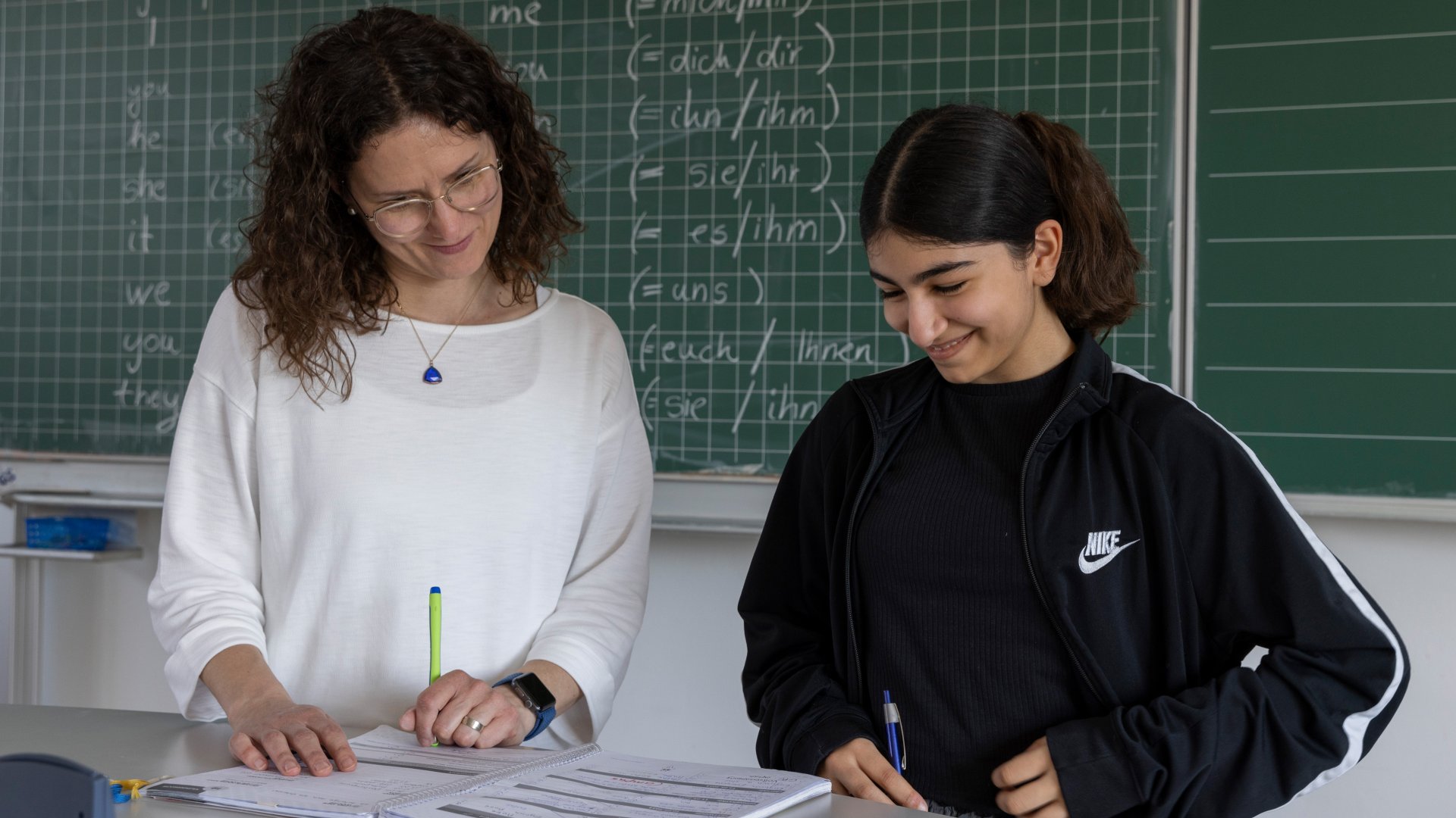A pupil and a teacher in conversation at Erlangen secondary school. Both are laughing. A blackboard can be seen in the background.