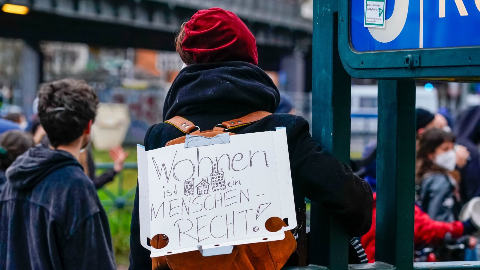 A woman looks into the distance, a sign is attached to her rucksack with the inscription: Housing is a human right