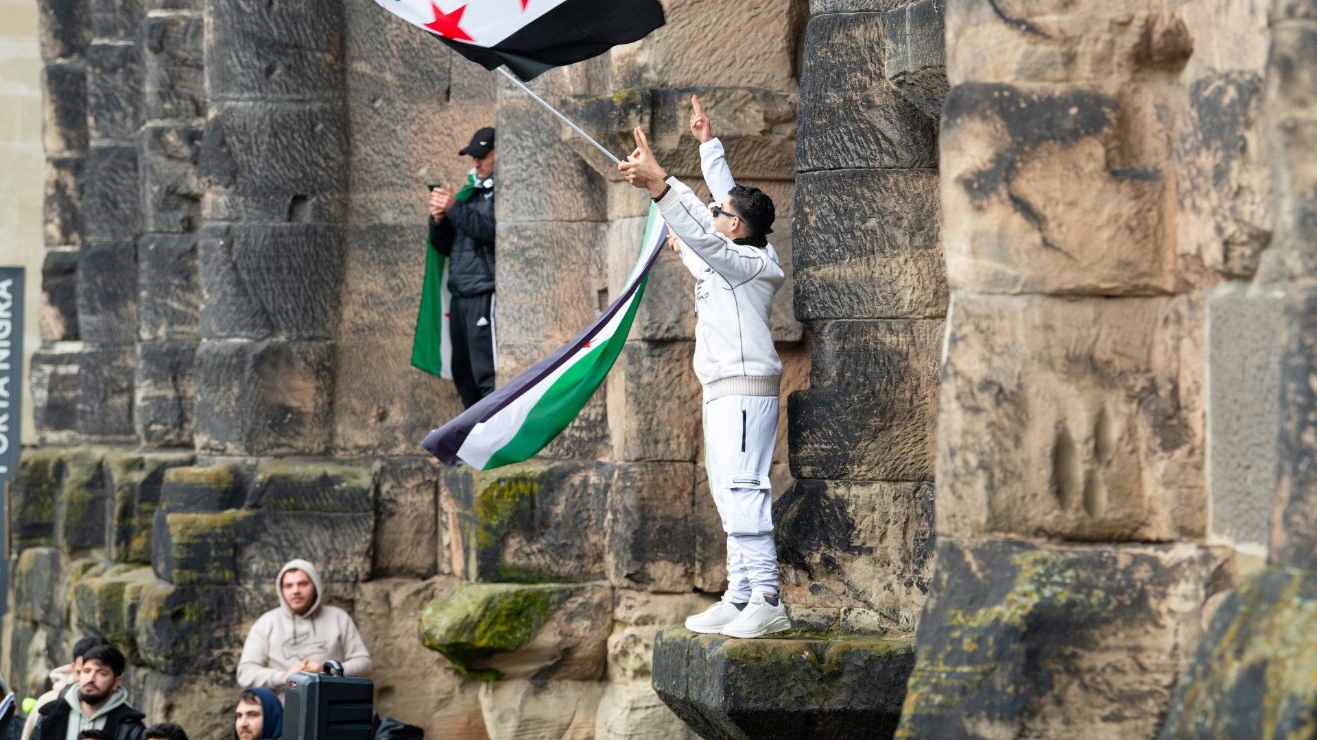 A man stands on a wall and waves the Syrian flag.