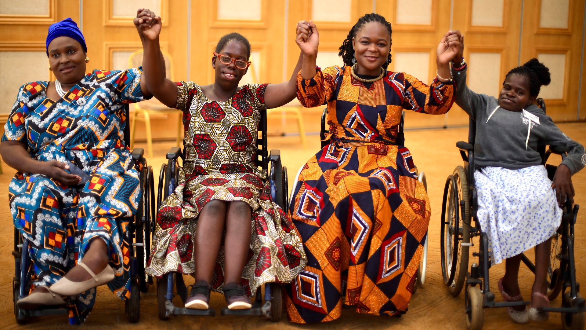 Four women from Malawi sit next to each other in wheelchairs, holding hands.